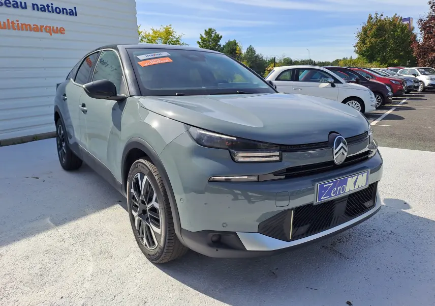 Vue 3/4 avant d'une Citroën C4 hybride 2025 Manhattan Green avec toit noir, sur parking extérieur ensoleillé.