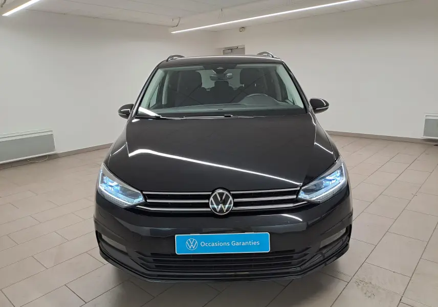 Vue avant d'un Volkswagen Touran 2025 noir grenadille avec phares allumés dans un showroom lumineux.