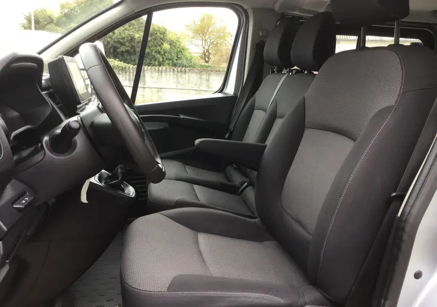 Vue intérieure côté conducteur du Renault Trafic Combi 2022, sièges tissu gris foncé avec accoudoir et volant multifonction.