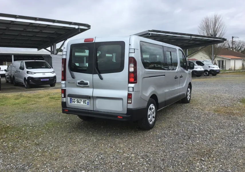 Vue 3/4 arrière droite d’un Renault Trafic Combi gris clair métal, avec portes arrière vitrées et plaque d’immatriculation française.
