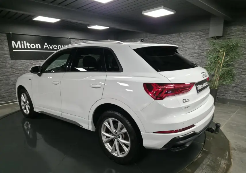 Audi Q3 blanc en 3/4 arrière droit, avec jantes S line et attelage amovible visible, dans un showroom intérieur.