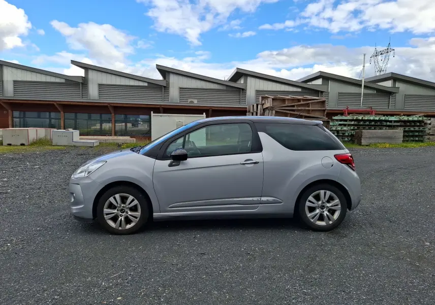 Profil côté gauche d'une Citroën DS3 gris clair avec toit noir et jantes alliage 16 pouces sur sol gravillonné.