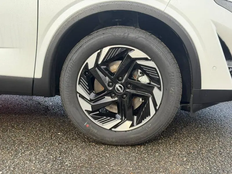Gros plan sur la roue avant gauche du Nissan Qashqai Hybrid e-POWER 2025 en blanc nacré avec jante noire et argentée au design moderne.