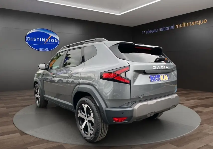 Vue 3/4 arrière droite d'un Dacia Duster gris foncé avec jantes alliage et barres de toit noires en showroom.