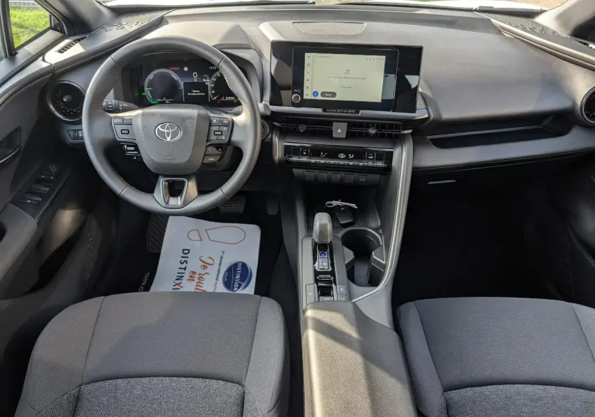Vue intérieure frontale du Toyota C-HR 2024, tableau de bord et volant avec écran tactile central et levier de boîte automatique.
