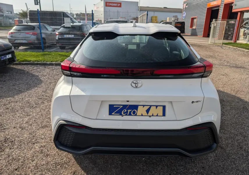 Vue arrière d'un Toyota C-HR blanc 2024 hybride, avec feux arrière en bandeau rouge et plaque d'immatriculation Zero KM.