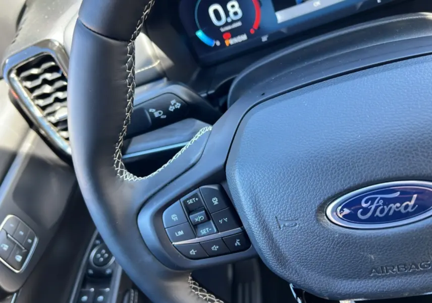 Gros plan sur le volant cuir multifonctions noir avec logo Ford et commandes intégrées, tableau de bord numérique en arrière-plan.
