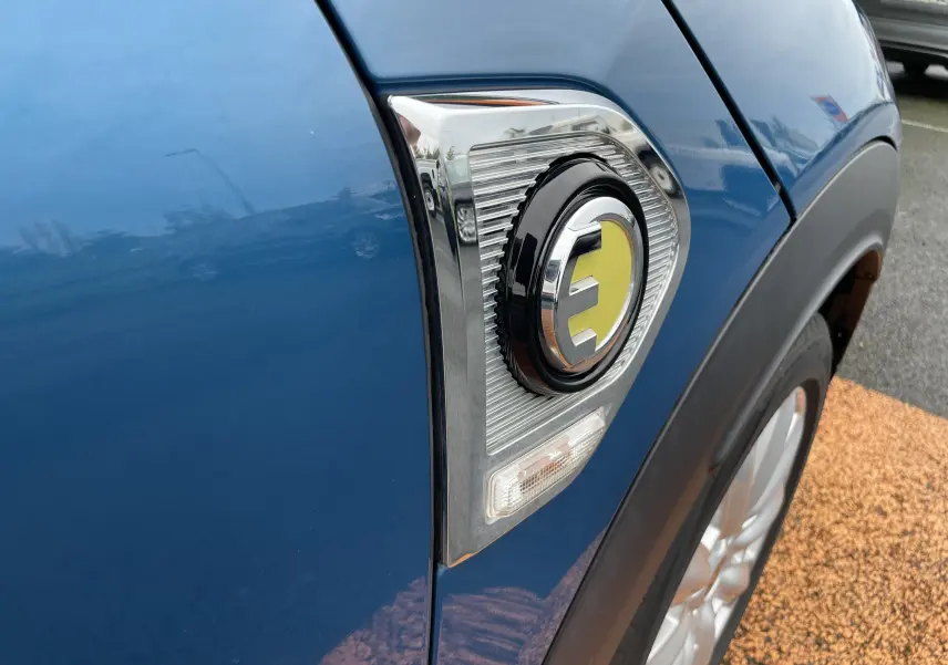 Gros plan sur l'emblème "E" jaune côté avant droit du MINI Countryman Cooper S bleu Island Metal hybride rechargeable.