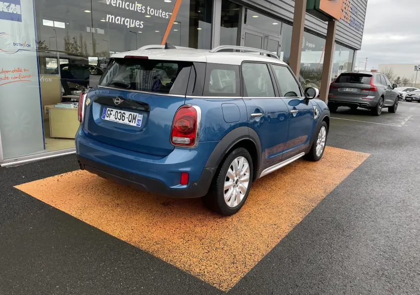 Vue 3/4 arrière droite d'une MINI Countryman Cooper S bleu Island Metal avec toit blanc, stationnée devant un showroom.
