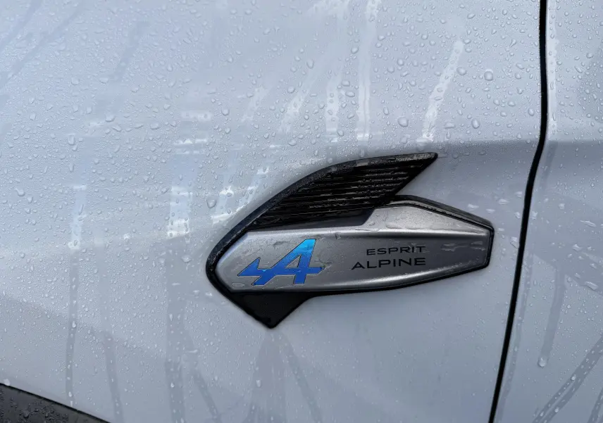 Détail du logo Esprit Alpine bleu sur la portière grise rafale d'une Renault Symbioz 2025, avec gouttes de pluie.