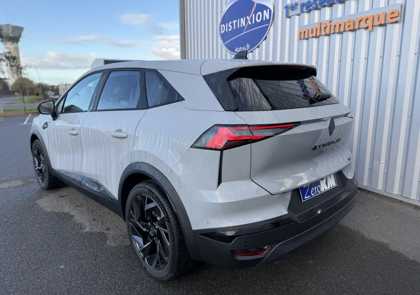 Vue 3/4 arrière droite du Renault SYMBIOZ gris rafale avec feux arrière LED et jantes noires, stationné devant un mur multimarque.