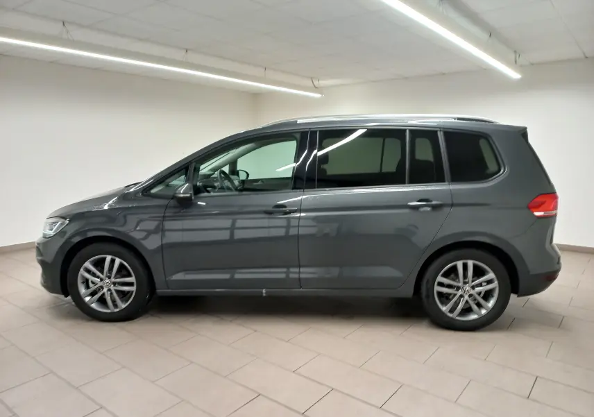 Profil côté gauche d'un Volkswagen Touran 2025 gris dauphin métallisé, version 7 places, en intérieur lumineux.