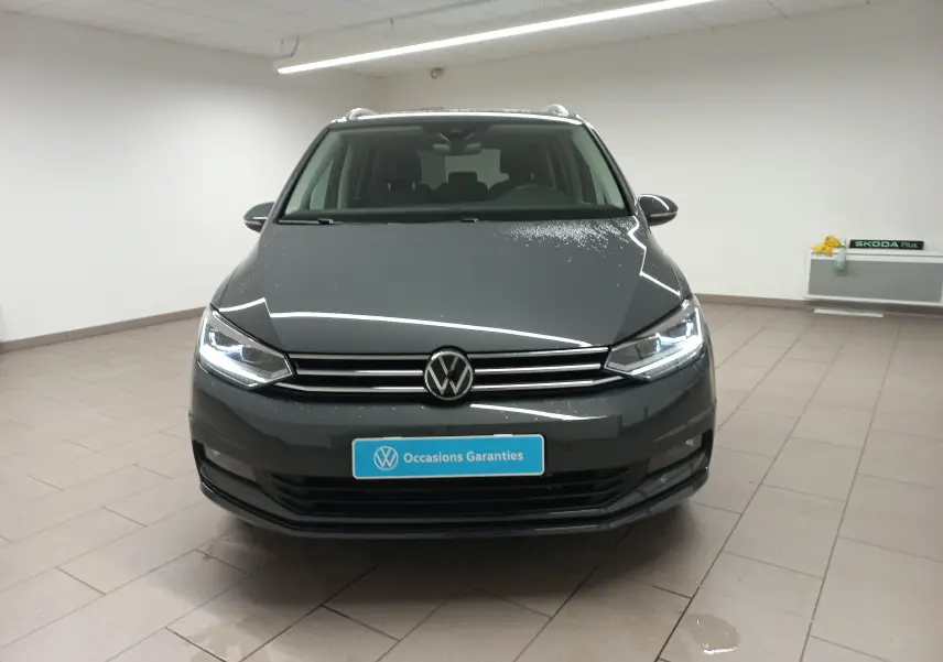 Vue frontale d'un Volkswagen Touran gris dauphin métallisé, avec phares allumés dans un showroom lumineux.