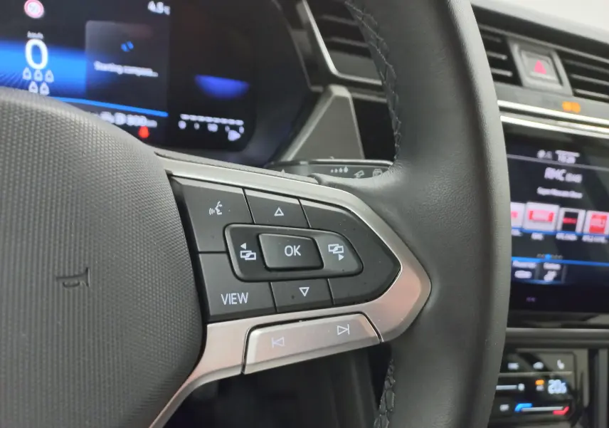 Gros plan sur les commandes au volant du Volkswagen Touran 2025, avec tableau de bord numérique et écran tactile en arrière-plan.