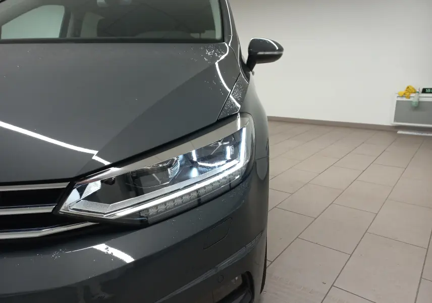 Gros plan sur le phare avant gauche du Volkswagen Touran 2025 gris dauphin avec peinture métallisée, en intérieur.