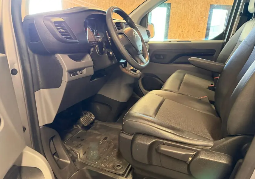 Vue intérieure côté conducteur du Citroën Jumpy gris 2022, montrant le volant, tableau de bord et sièges avant en tissu noir.