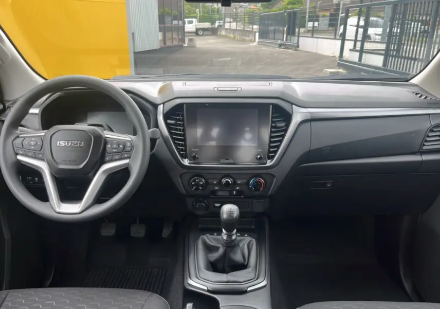 Vue intérieure avant de l'Isuzu D-MAX 2026 avec tableau de bord noir, volant multifonctions et boîte manuelle six vitesses.