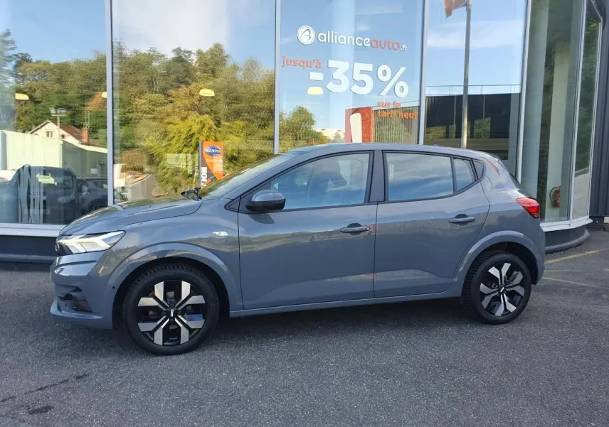 Profil côté gauche d'une Dacia Sandero 2024 gris urbain avec jantes alu 16 pouces, garée devant une vitrine.