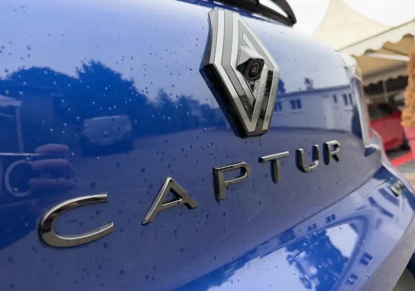 Gros plan sur le logo Renault et le nom Captur en lettres chromées sur la malle arrière bleu brillant avec gouttes d'eau.