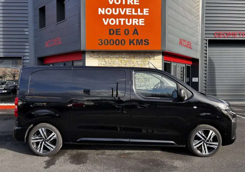 Profil droit d’un FIAT Scudo Fourgon noir 2025, avec jantes alliage et vitres teintées, stationné devant un bâtiment commercial.