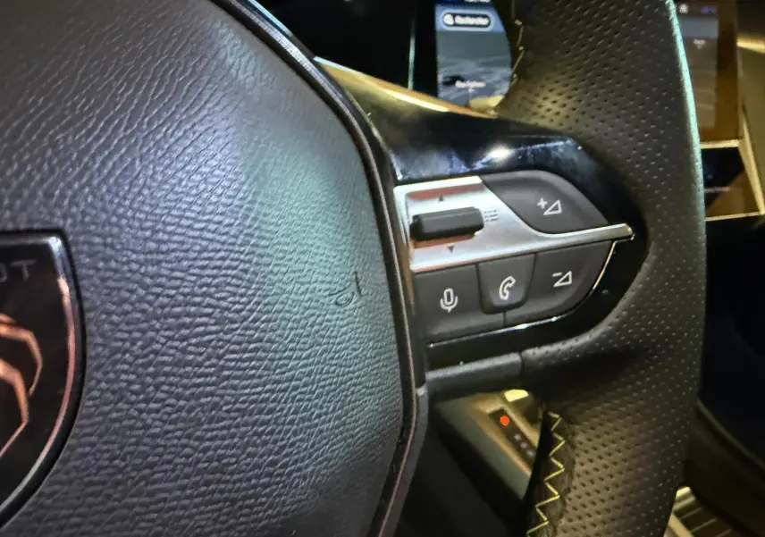 Gros plan sur le volant noir perforé avec commandes intégrées et écran tactile flou en arrière-plan.