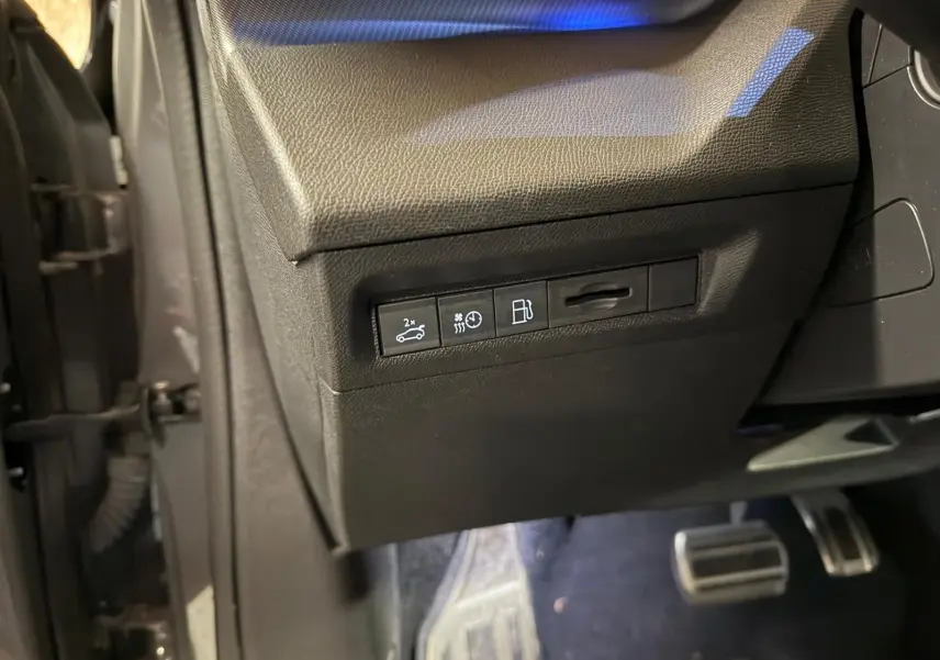 Vue intérieure côté conducteur du tableau de bord noir avec commandes d'ouverture de coffre et réglages, pédales aluminium visibles.