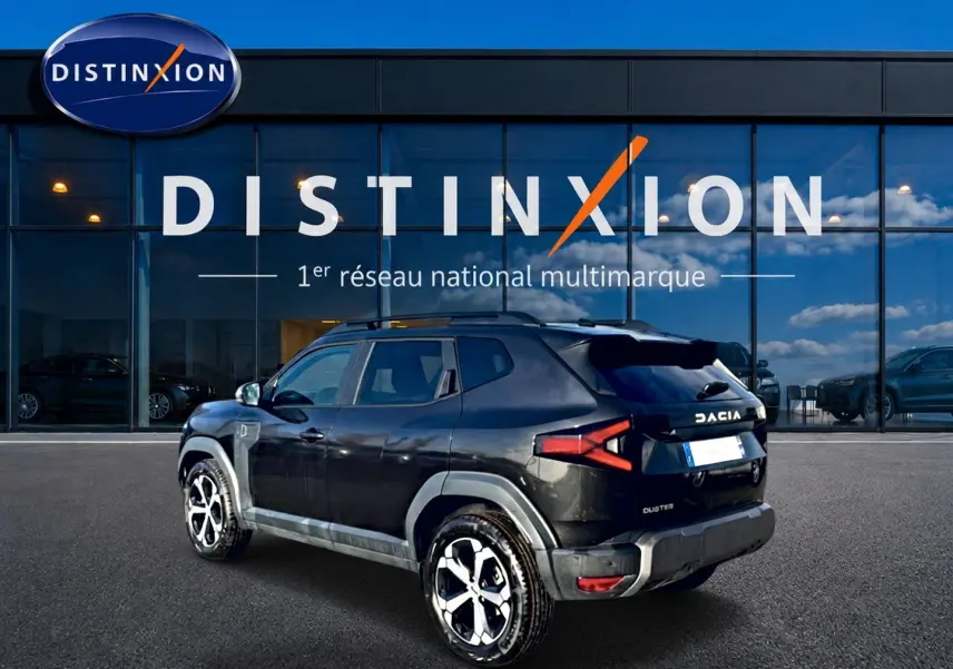 Vue 3/4 arrière droite du Dacia Duster noir 2026 avec jantes alu et barres de toit devant un showroom Distinxion.