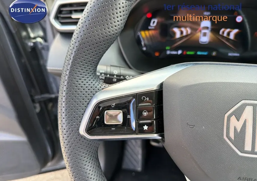 Gros plan sur le volant perforé gris du MG ZS 1.5L Hybrid+ 2025, avec commandes intégrées et tableau de bord numérique en arrière-plan.