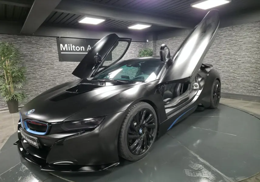 BMW i8 Coupé Pure Impulse noir mat vue 3/4 avant droit avec portes en élytre ouvertes dans un showroom.