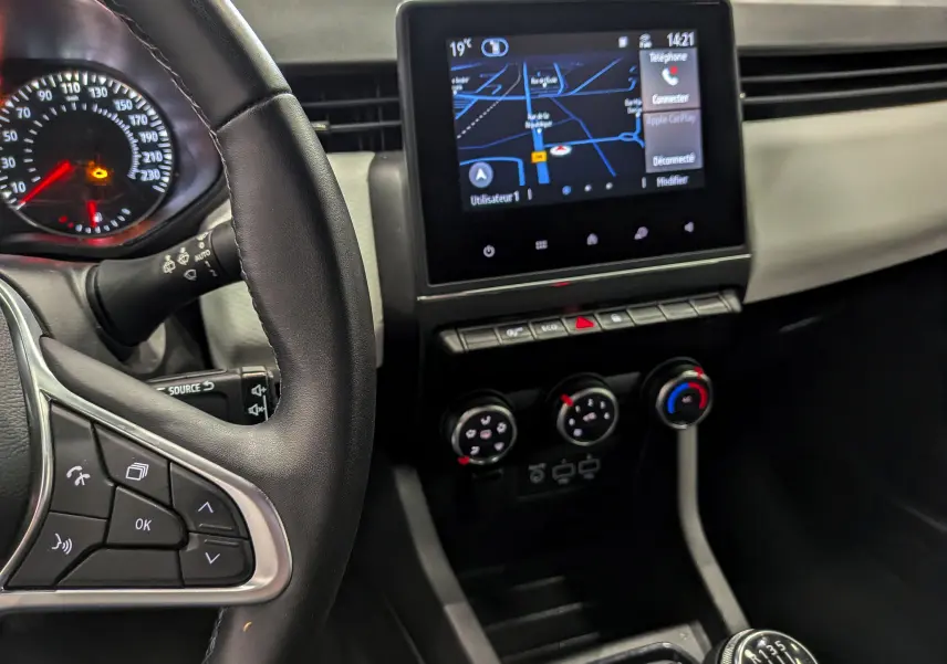 Vue rapprochée de l’intérieur de la Renault Clio V blanc glacier, montrant le volant, l’écran tactile avec GPS et la boîte manuelle.