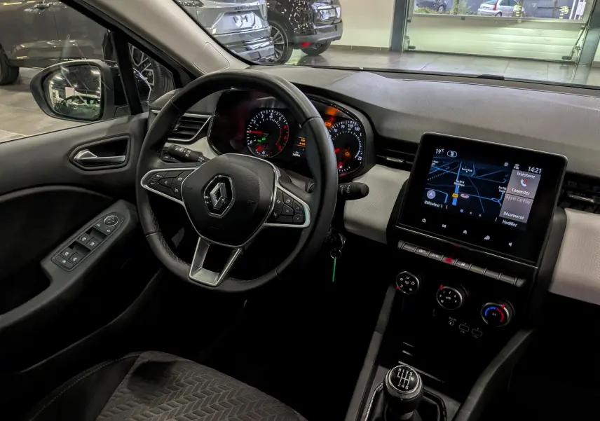 Vue intérieure côté conducteur de la Renault Clio V blanc glacier, avec tableau de bord, volant multifonction et écran tactile central.
