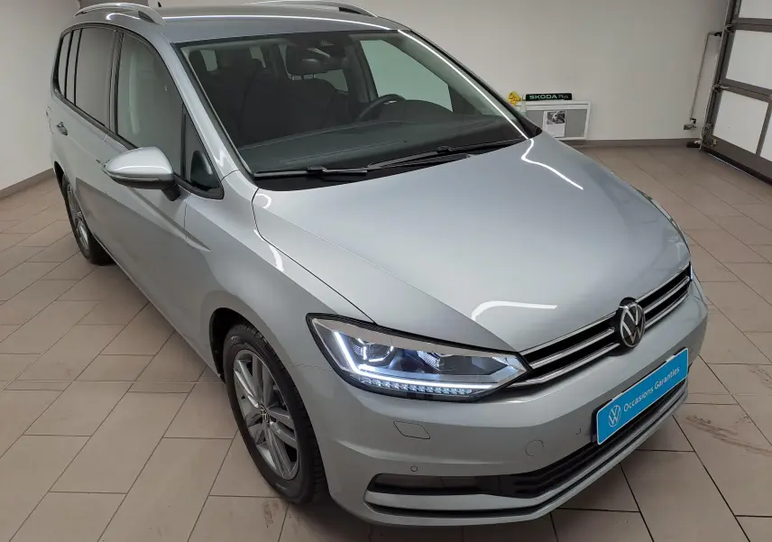 Vue 3/4 avant droit d'un Volkswagen Touran 2025 argent Dolomite avec phares LED allumés en intérieur showroom.