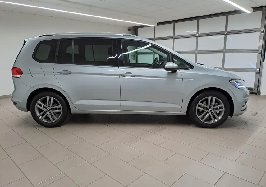 Profil droit d'un Volkswagen Touran 2025 argent Dolomite avec jantes alliage et vitres teintées, en intérieur showroom.
