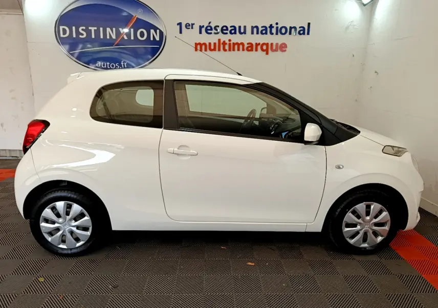 Profil côté droit d'une Citroën C1 blanche 3 portes, avec jantes acier et enjoliveurs, en intérieur showroom.