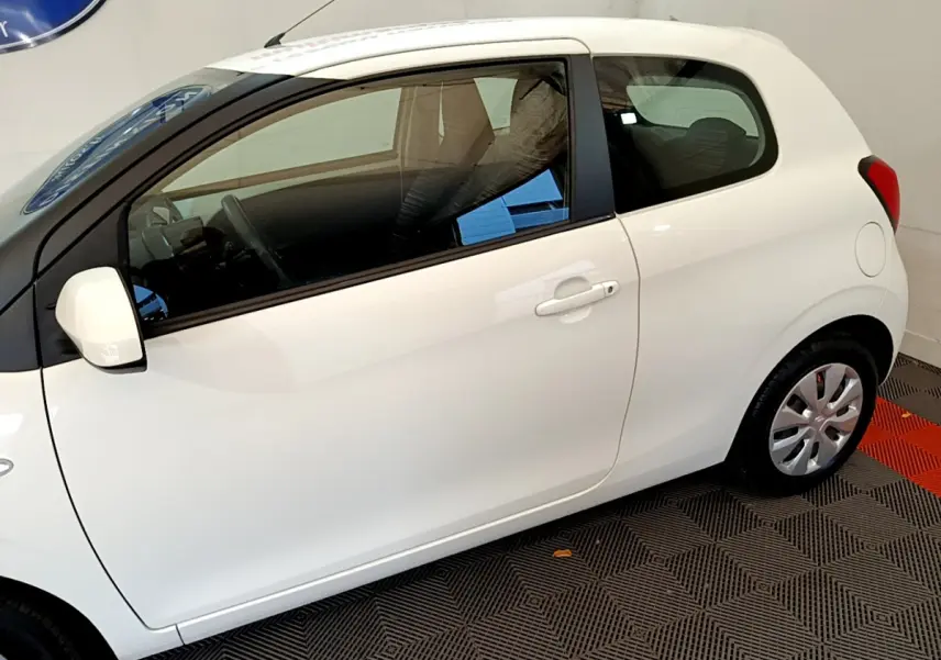 Profil droit d'une Citroën C1 blanche 3 portes, avec rétroviseur et jante acier visibles, sur sol intérieur.