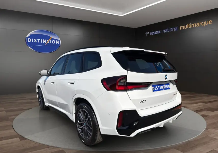 BMW X1 sDrive20d blanc vue 3/4 arrière droit, avec jantes noires et aileron de toit visible.