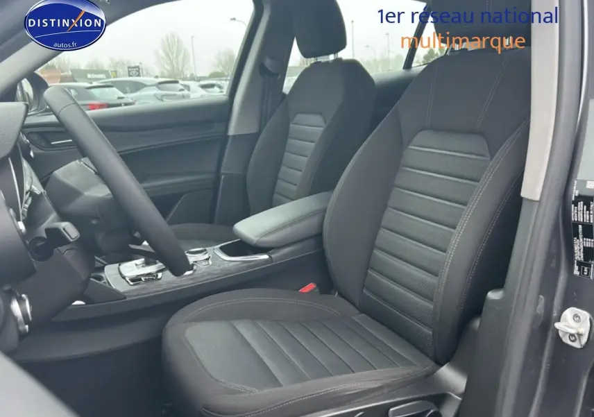 Vue intérieure côté conducteur de l'Alfa Romeo Stelvio gris Vesuvio, mettant en valeur les sièges et l'accoudoir central.