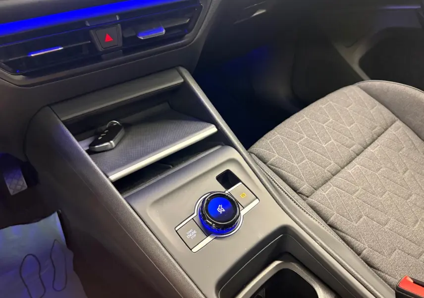 Gros plan sur la console centrale du Volkswagen Tiguan 2025, avec molette de commande éclairée en bleu et siège tissu gris.