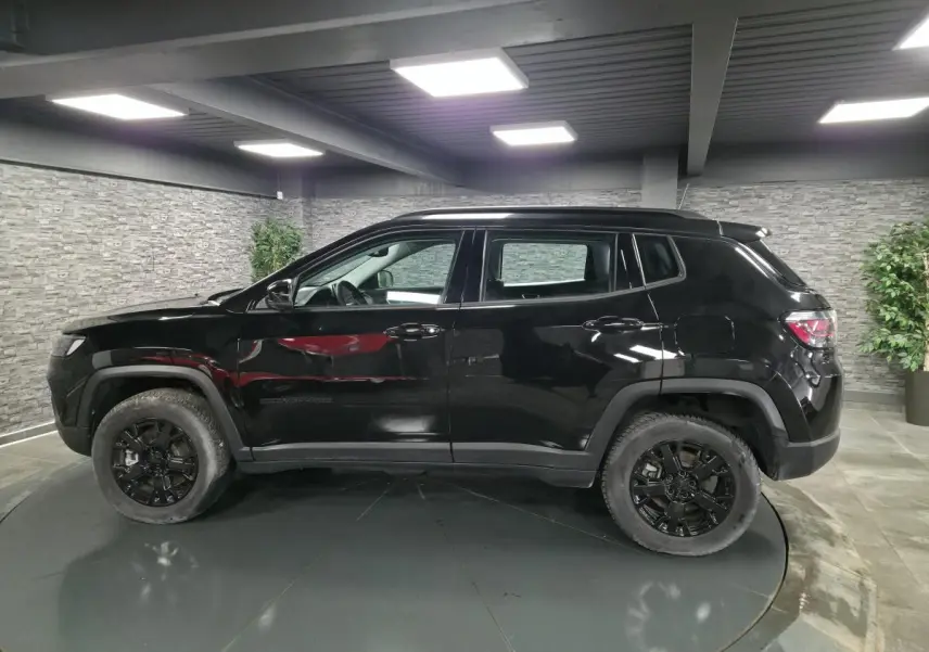Vue de profil côté gauche d'un Jeep Compass noir avec jantes noires et garde au sol surélevée dans un showroom.