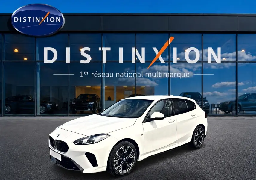 BMW Série 1 blanche vue en 3/4 avant droit, avec jantes alliage noires et calandre M Sport devant un showroom Distinxion.