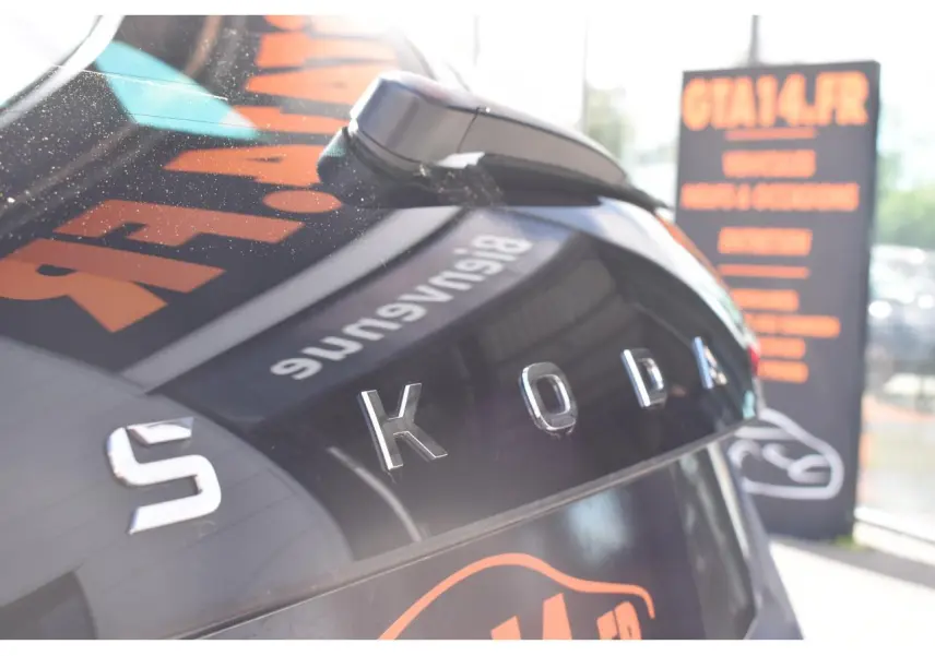 Gros plan sur l’arrière gris graphite d’une Skoda Scala 2025 avec logo SKODA en relief et reflet d’enseigne orange.
