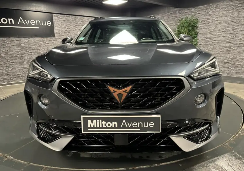 Vue frontale d'un CUPRA Formentor gris magnétique avec calandre noire et logo cuivre, dans un showroom intérieur.