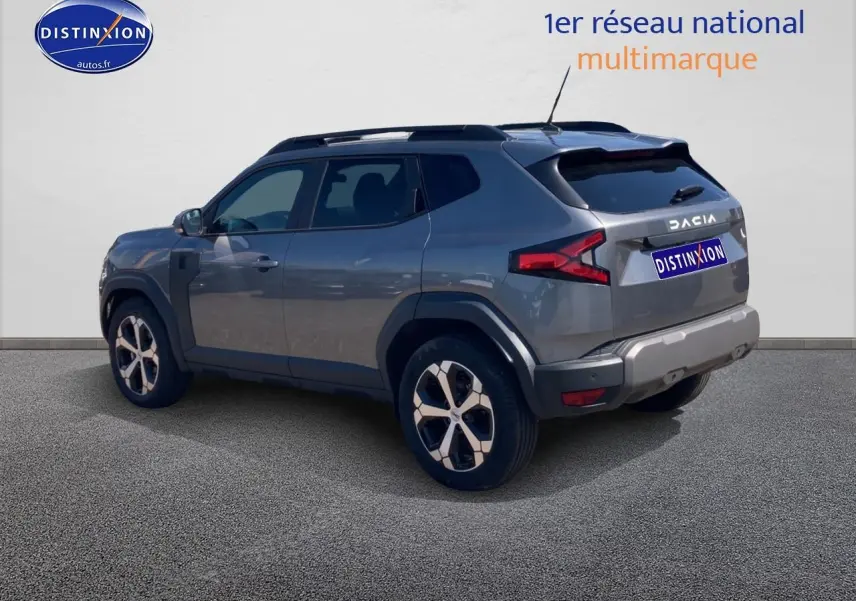 Vue 3/4 arrière droite d'un Dacia Duster gris schiste avec barres de toit et jantes alu distinctives.