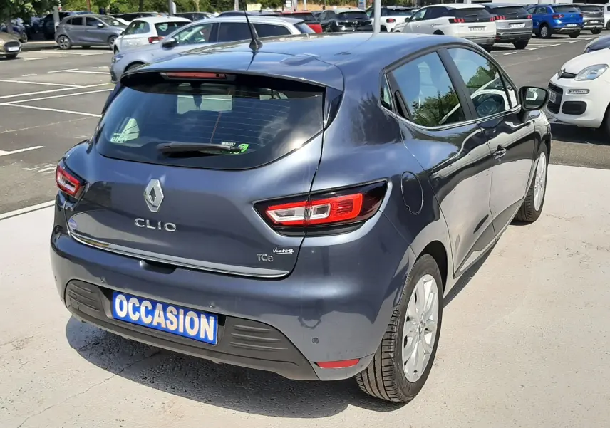 Vue 3/4 arrière droite d'une Renault Clio IV gris titanium, avec plaque "OCCASION" sur un parking extérieur.