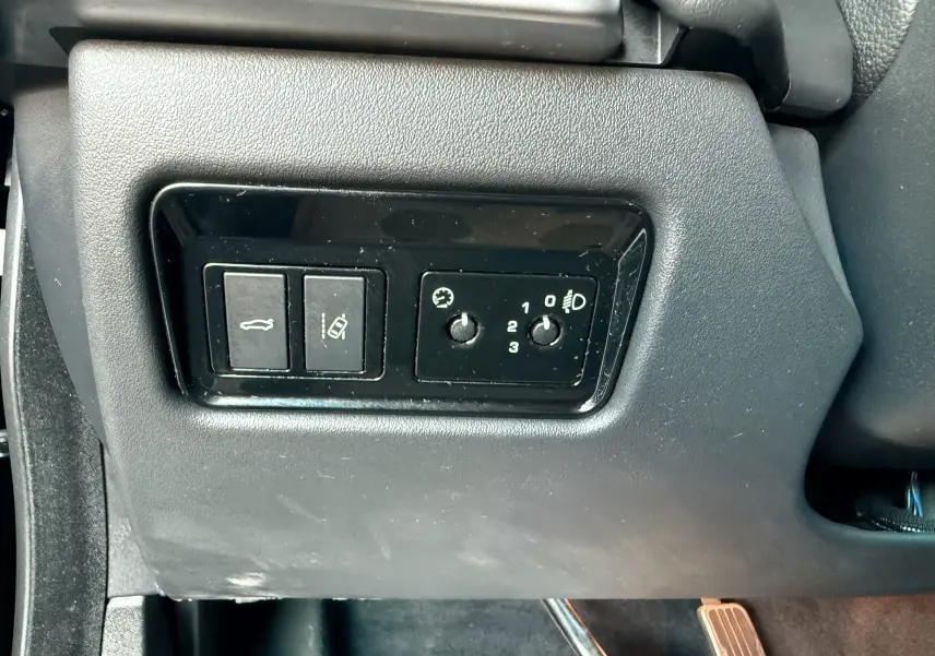 Gros plan sur les commandes de réglage des phares et ouverture du coffre côté gauche dans l'habitacle noir d'une Jaguar XE 2019.