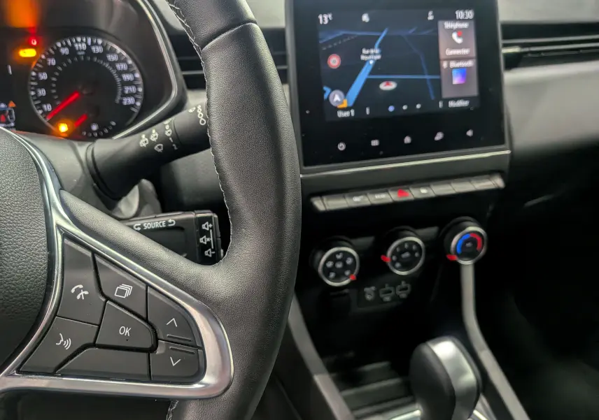 Intérieur de la Renault Clio V blanc glacier, vue rapprochée du volant à gauche et de l’écran tactile central avec navigation.