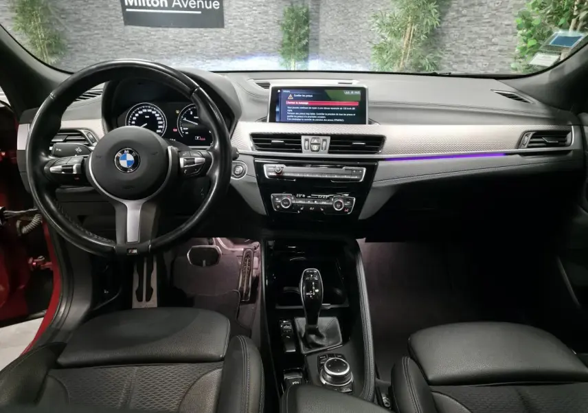 Vue intérieure avant de la BMW X2 orange, tableau de bord avec éclairage d'ambiance violet et volant M cuir noir.