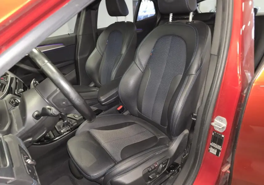 Vue intérieure côté conducteur de la BMW X2 orange 2021, sièges sport en cuir noir avec surpiqûres bleues et éclairage d'ambiance violet.