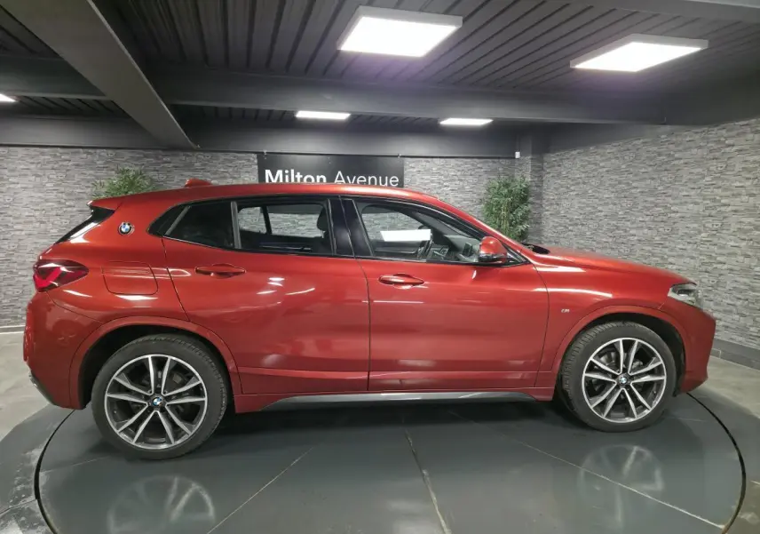 Vue de profil côté gauche d'un BMW X2 orange métallisé avec jantes alliage 19 pouces et badge M Sport visible.