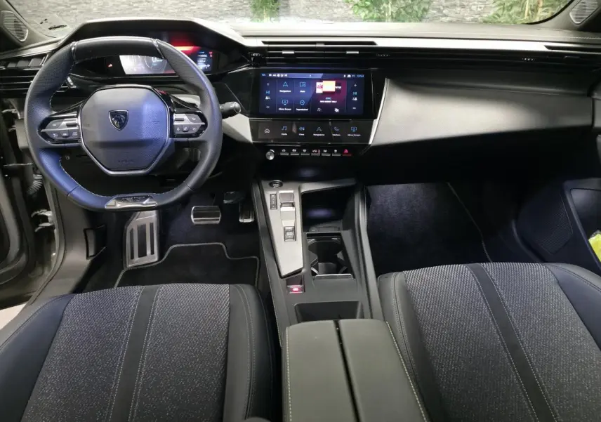 Intérieur Peugeot 408 GT hybride 2025 vu de face, volant cuir perforé, écran tactile central et sièges tissu noir.